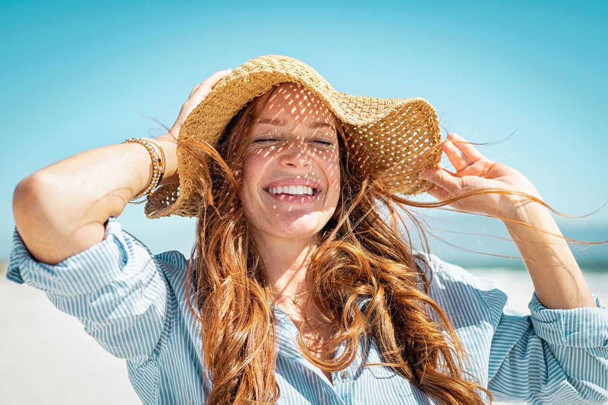 Smiling woman with freckles shading her face with a straw sun hat on a bright beach