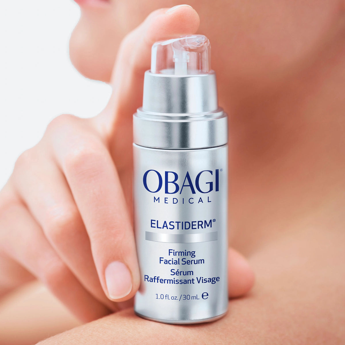 A close-up of a person holding a silver Obagi Medical Elastiderm facial serum bottle against a soft skin-toned background.