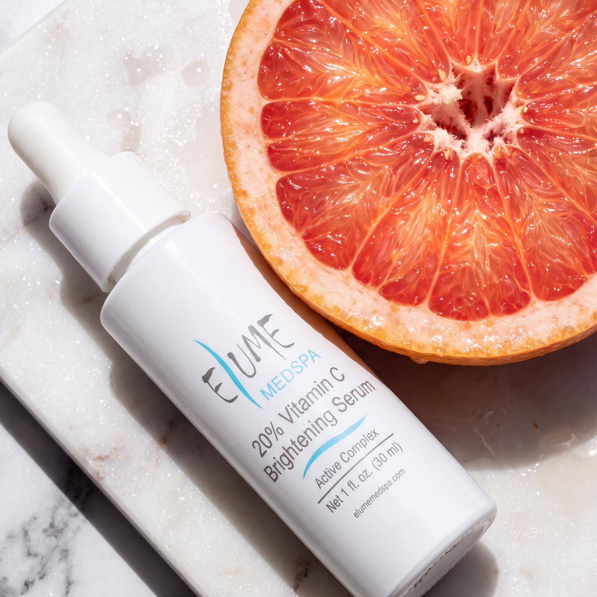 A white dropper bottle of Eume Medspa Brightening Serum placed diagonally on a white marble slab next to a sliced grapefruit.