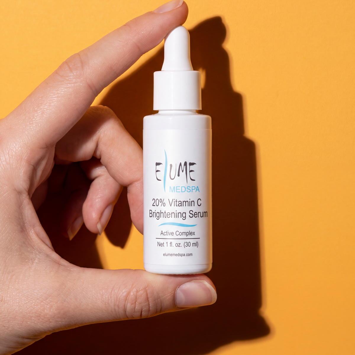 A hand holding a white dropper bottle of Elume Medspa 20% Vitamin C Brightening Serum against a solid orange background.
