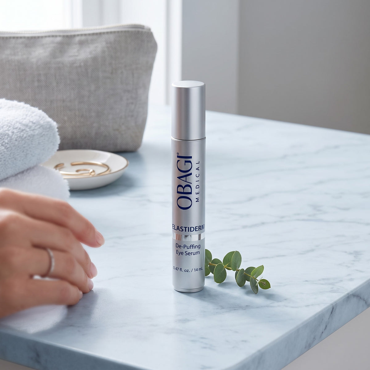 A silver bottle of OBAGI Elastiderm De-Puffing Eye Serum stands on a light marble countertop, with a hand resting nearby and a gray pouch in the background.
