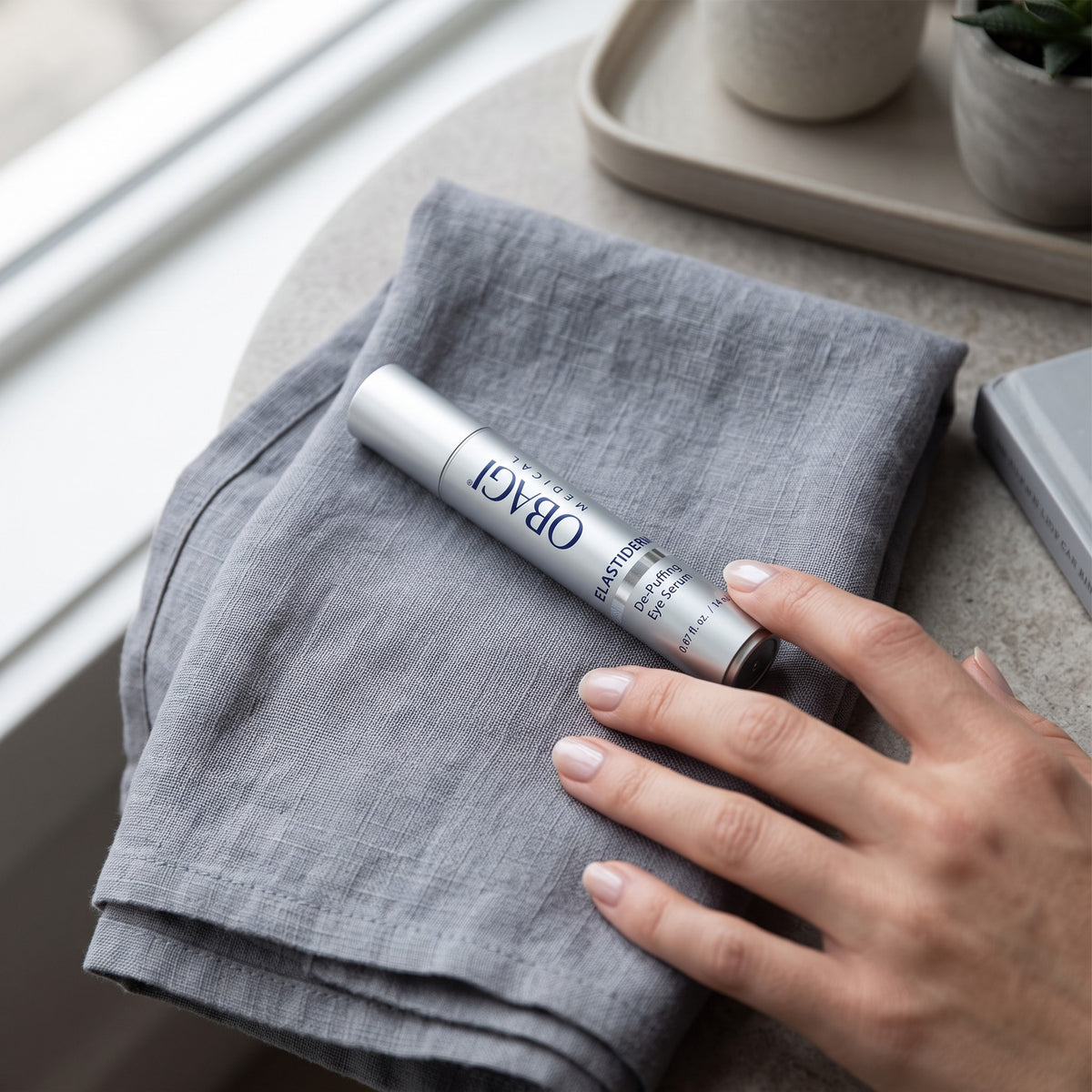 A hand rests on a folded gray linen cloth next to a silver roll-on bottle labeled ORACI HAND CARE, set on a stone surface near a window.