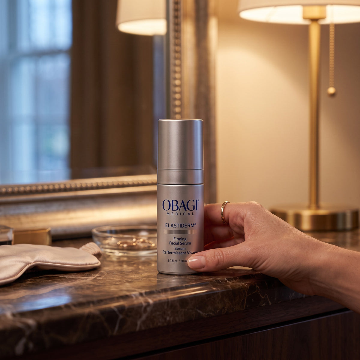 A hand holds a silver bottle of Obagi Medical Elastderm Firming Retinol Cream on a dark marble vanity countertop, with a mirror, gold-framed picture frame, and lit table lamps in the background.