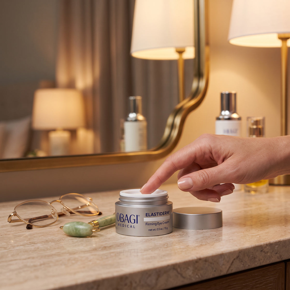 A hand reaches for the white lid of a silver and blue UBAGI MEDICAL Elastinica cream jar placed on a marble vanity counter, with eyeglasses, a jade roller, and a blurred bathroom mirror in the background.
