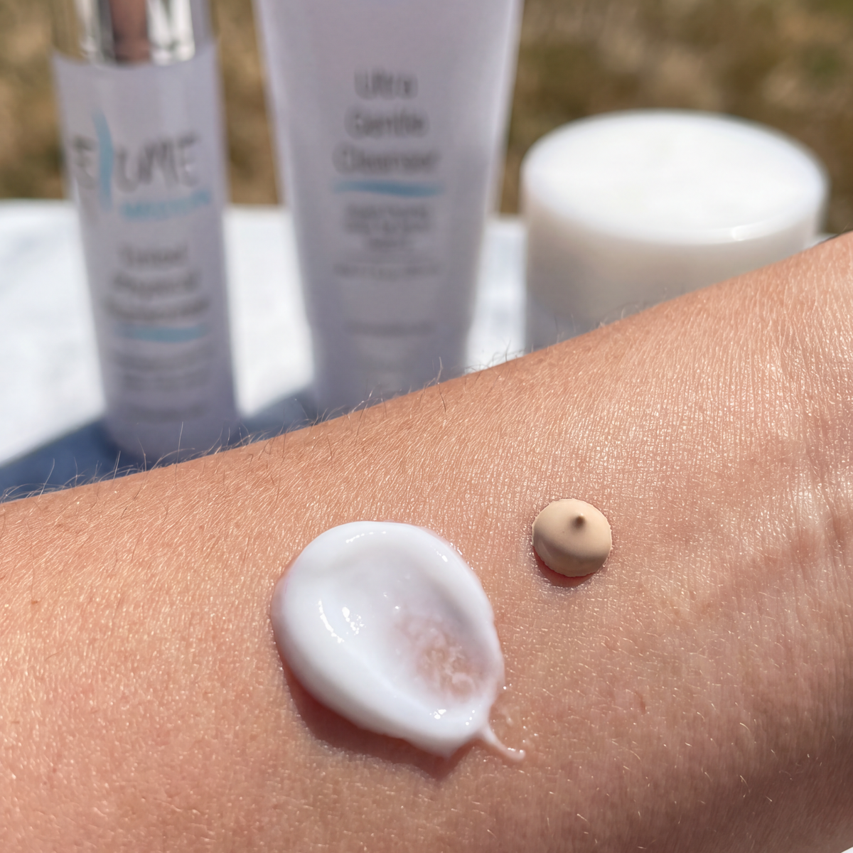 A close-up of a person's forearm showing a dollop of white cream and a small beige droplet, with two skincare bottles in the blurred background. The visible text on the bottles includes 'ELUME' and 'Medium'.
