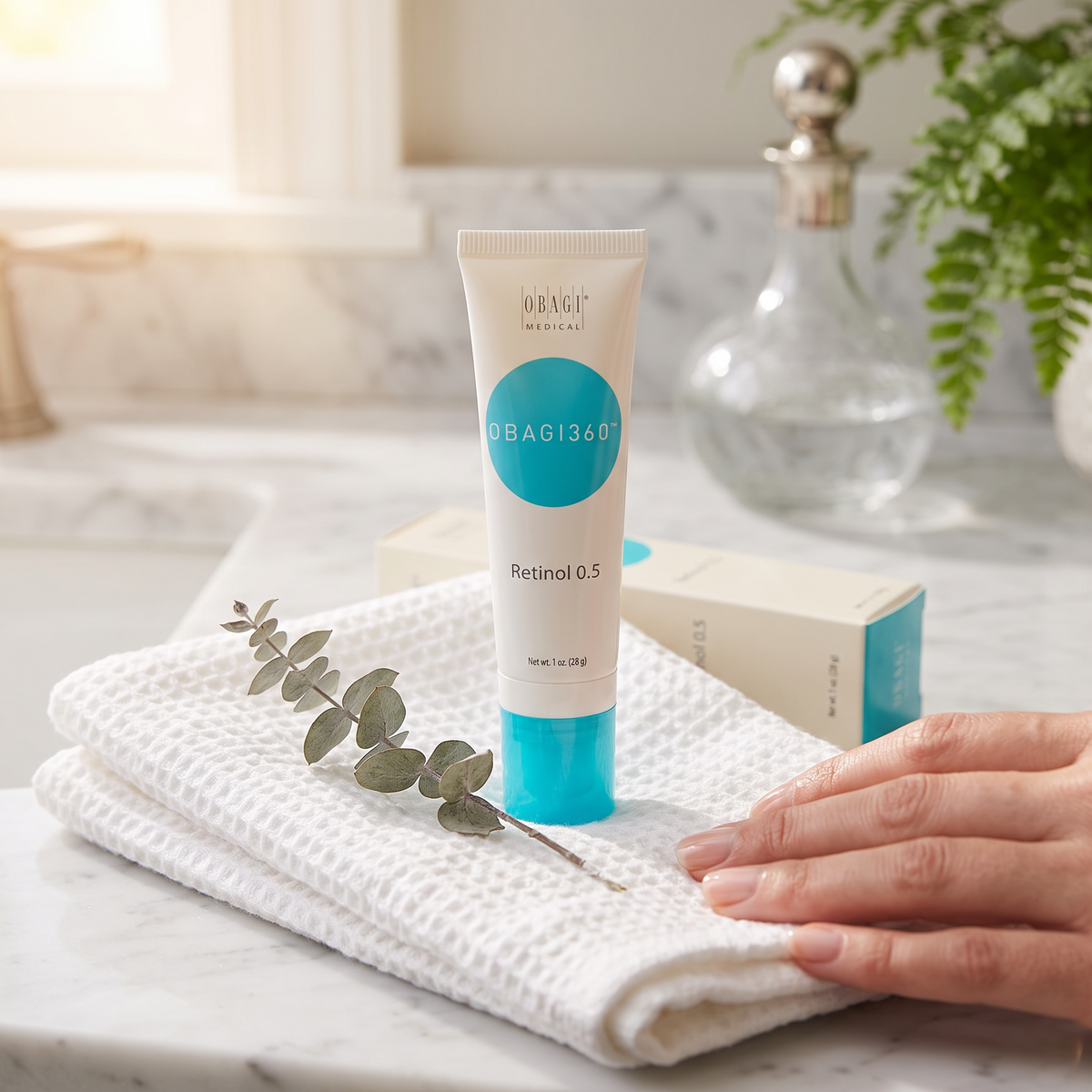 Elume Medspa Obagi360 Retinol 0.5 cream tube on a white towel with a sprig of eucalyptus on a marble countertop.