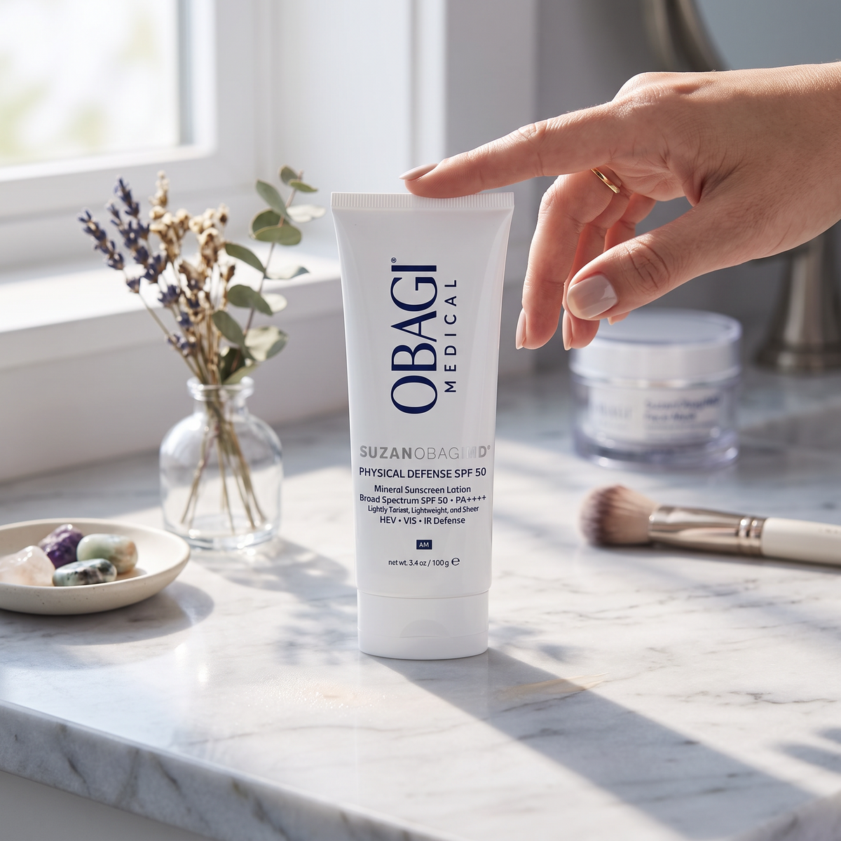 Elume Medspa SUZANOBAGIMD Tinted Physical Defense SPF 50 mineral sunscreen tube on a marble vanity with a hand.