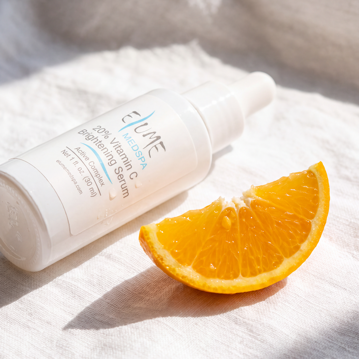 A white bottle of Elume 20% Vitamin C Brightening Serum lies on a white textured fabric next to a fresh orange slice, with bright natural sunlight casting soft shadows.