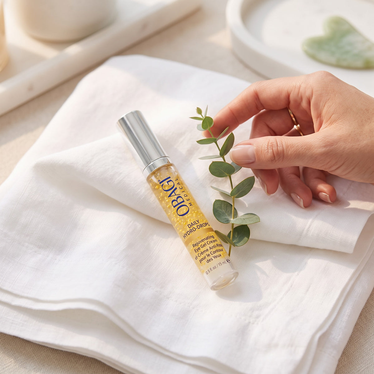 A clear cylindrical cosmetic roller bottle filled with golden-yellow oil, placed on a folded white towel. A hand with a gold ring gently holds a green eucalyptus stem beside it.