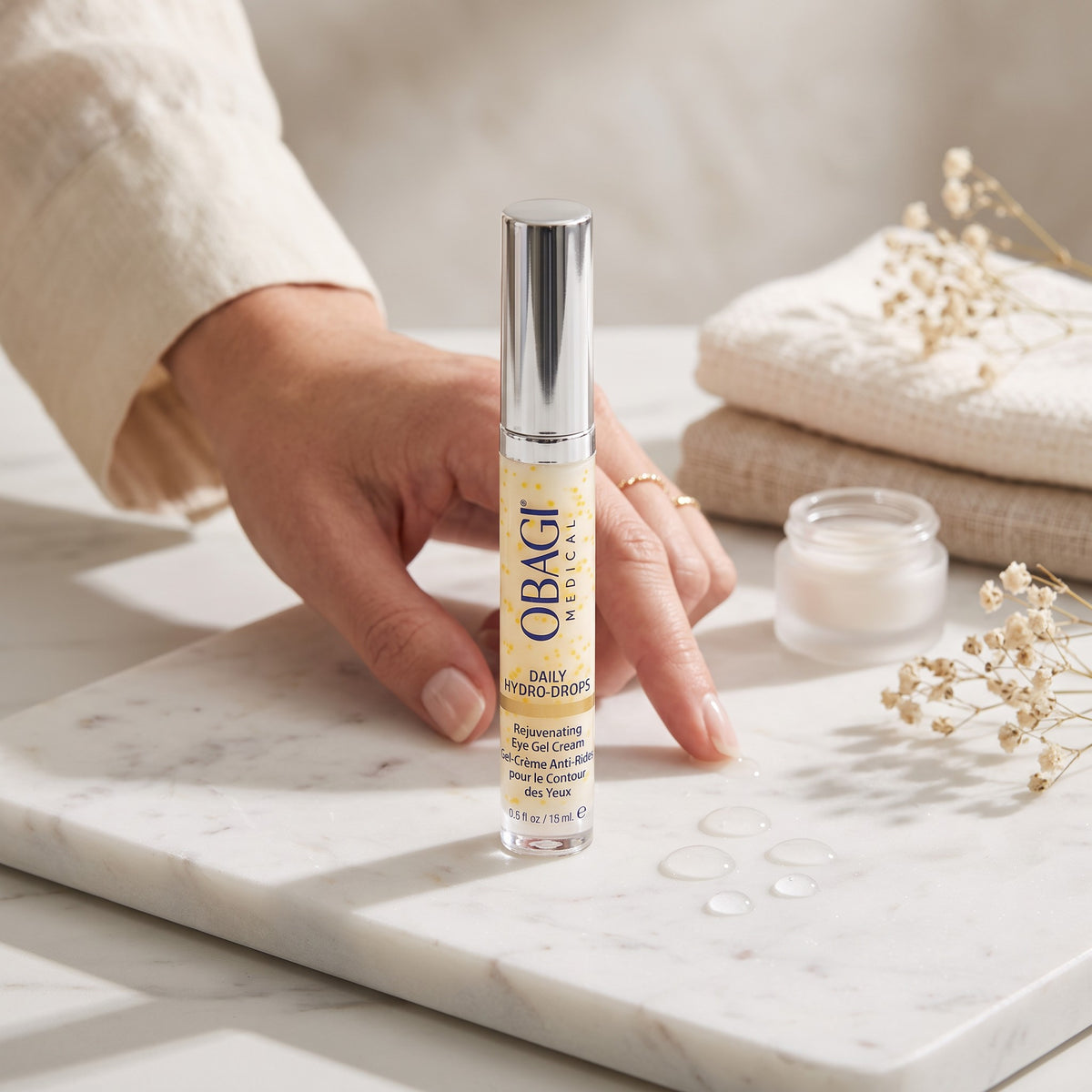 A hand holds a tube of Obagi Daily Hydro Cream against a styled background with a marble tray, folded cream towels, and dried flowers. The Obagi Daily Hydro Cream tube is centered, with the Obagi brand name clearly visible.