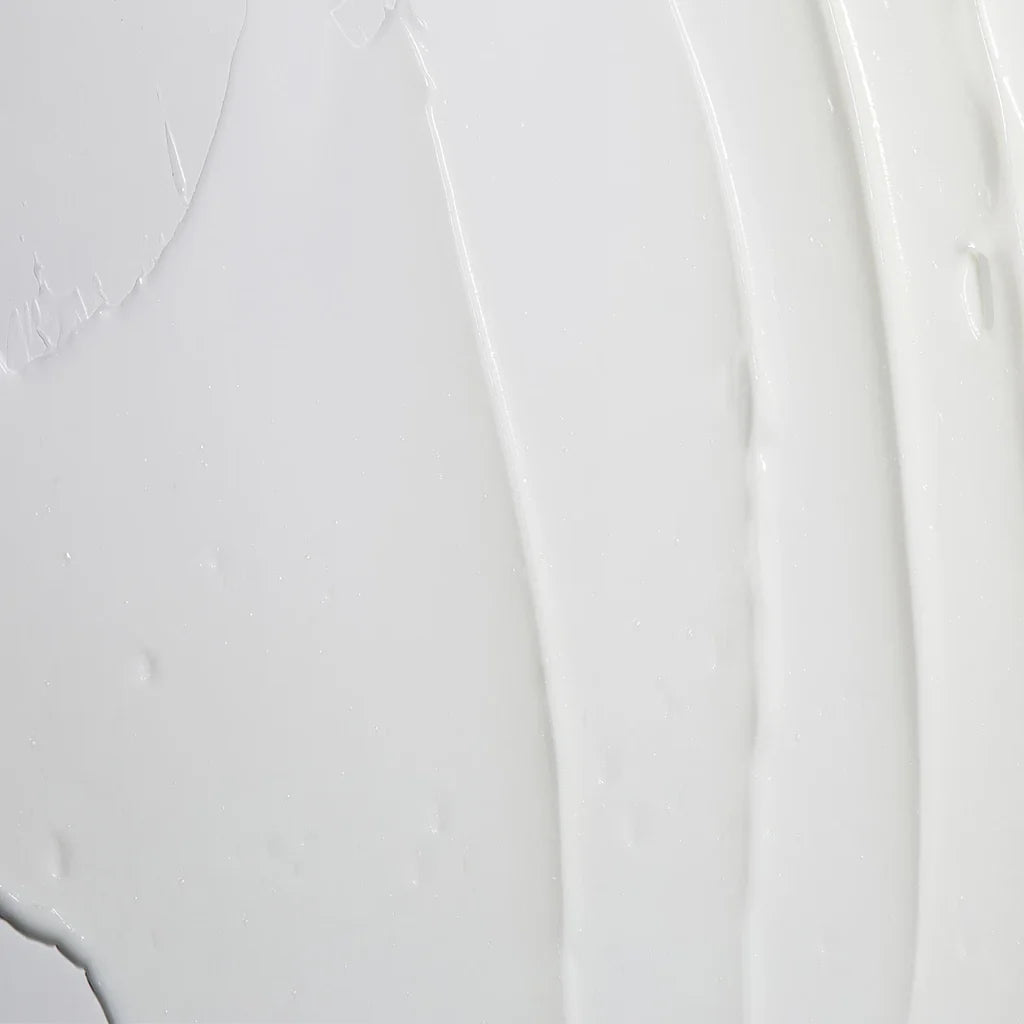 A close-up texture shot of a thick, creamy white cosmetic cream with smooth vertical swatches and small droplets on a light gray surface.