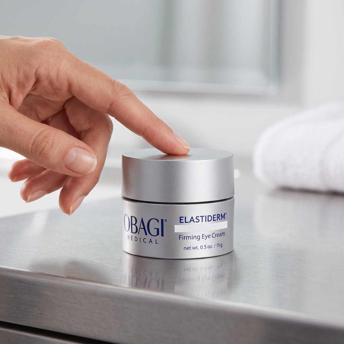 A hand gently resting on the lid of a silver jar of Obagi Medical Elastiderm Firming Eye Cream, placed on a reflective stainless steel counter with a blurred white towel in the background.