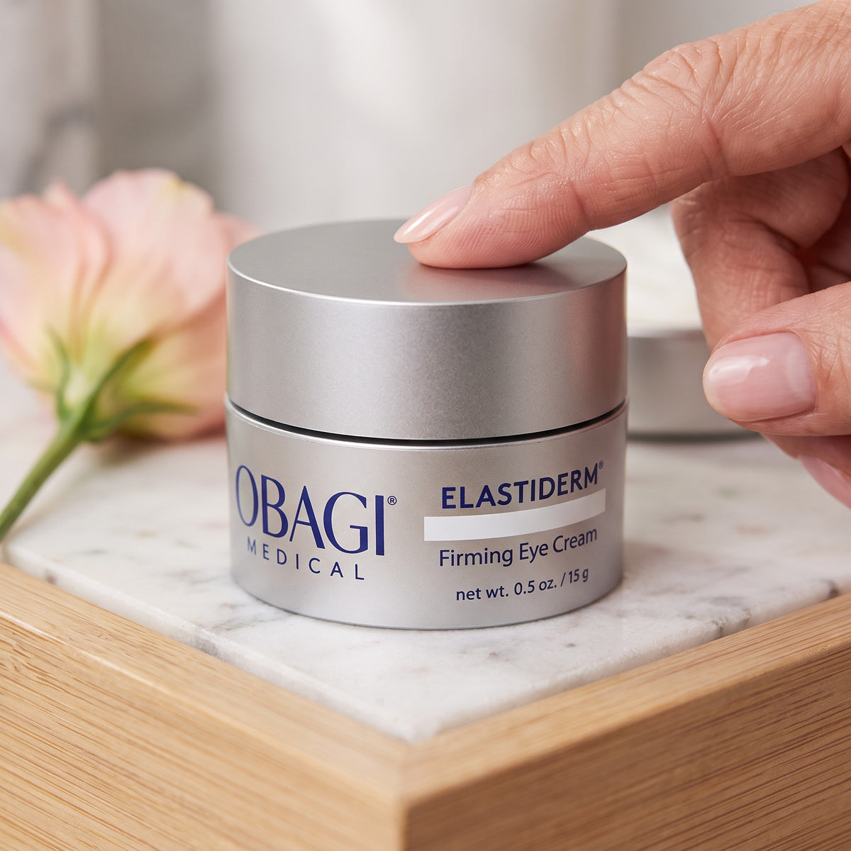 A close-up of a hand resting on the lid of an Obagi Medical Elastiderm Firming Eye Cream jar, placed on a marble tray with a soft pink flower in the background.