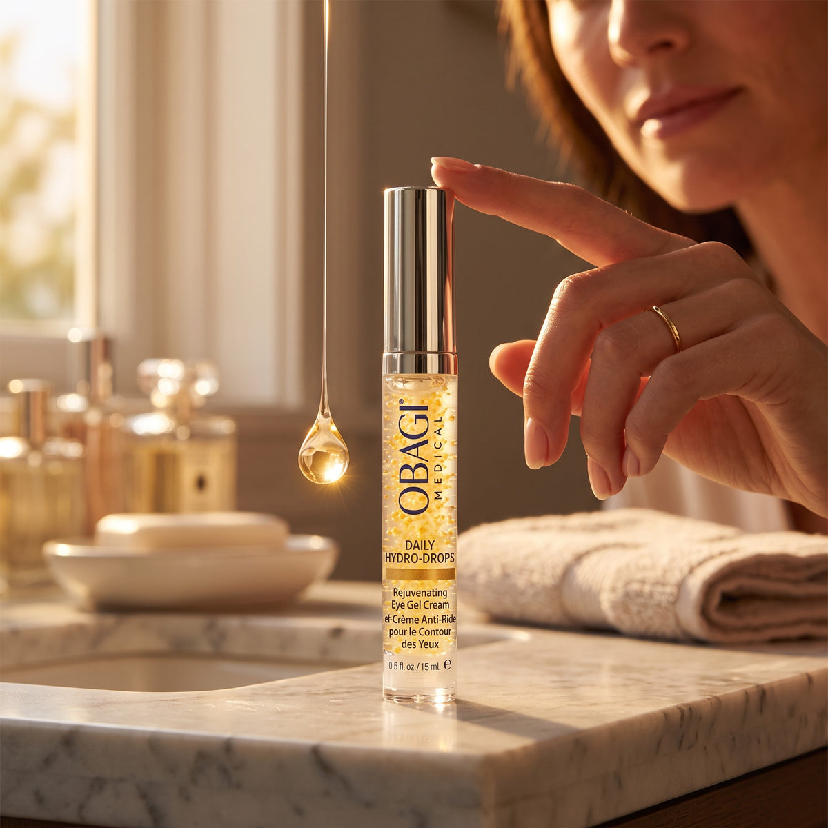 A close-up lifestyle shot of a woman's hand touching a bottle of OBAGI Daily Hydro Drops eye gel on a marble bathroom counter, with a warm, sunlit window and toiletries blurred in the background.