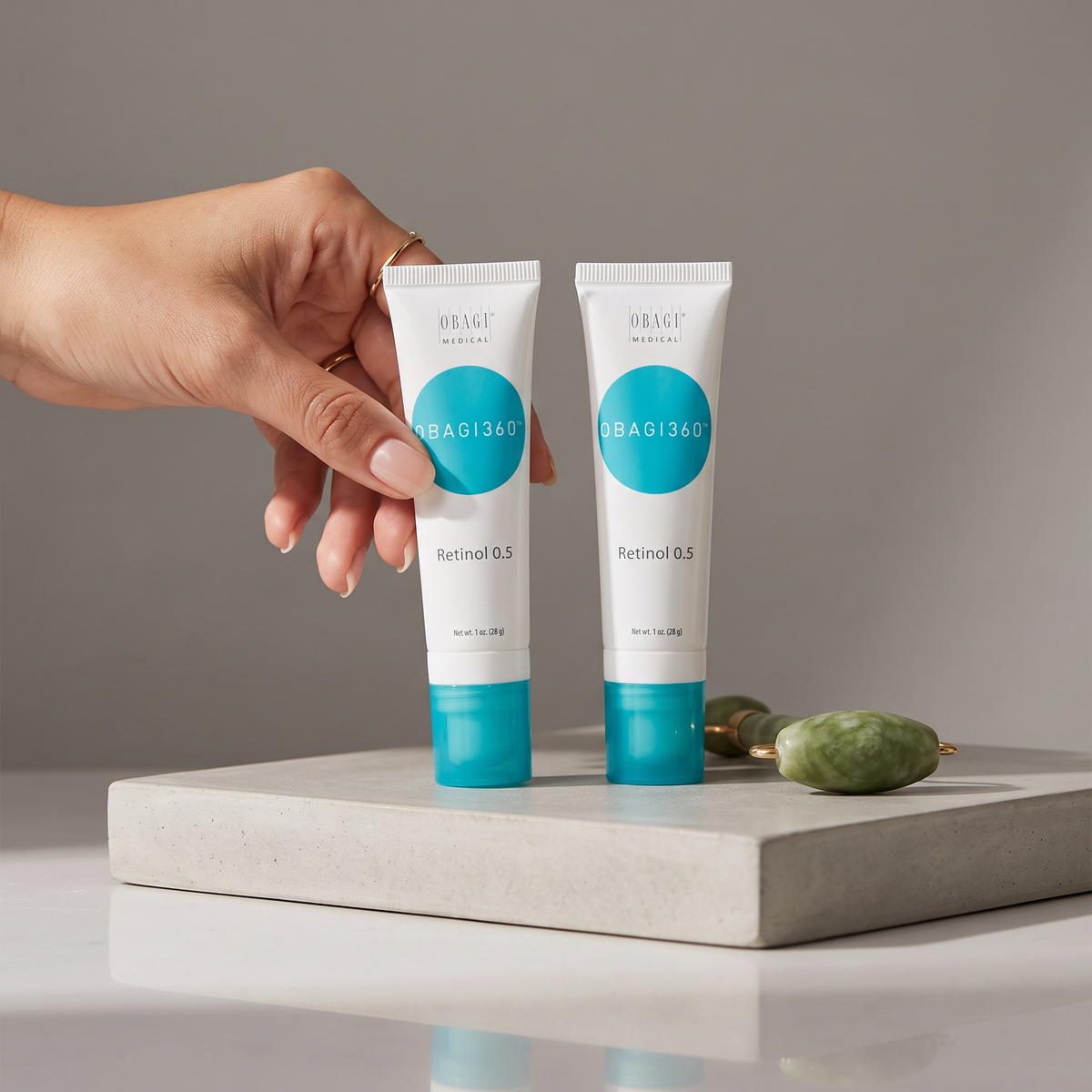 Elume Medspa Obagi360 Retinol 0.5 tubes held by a hand next to a green jade roller on a concrete display block.