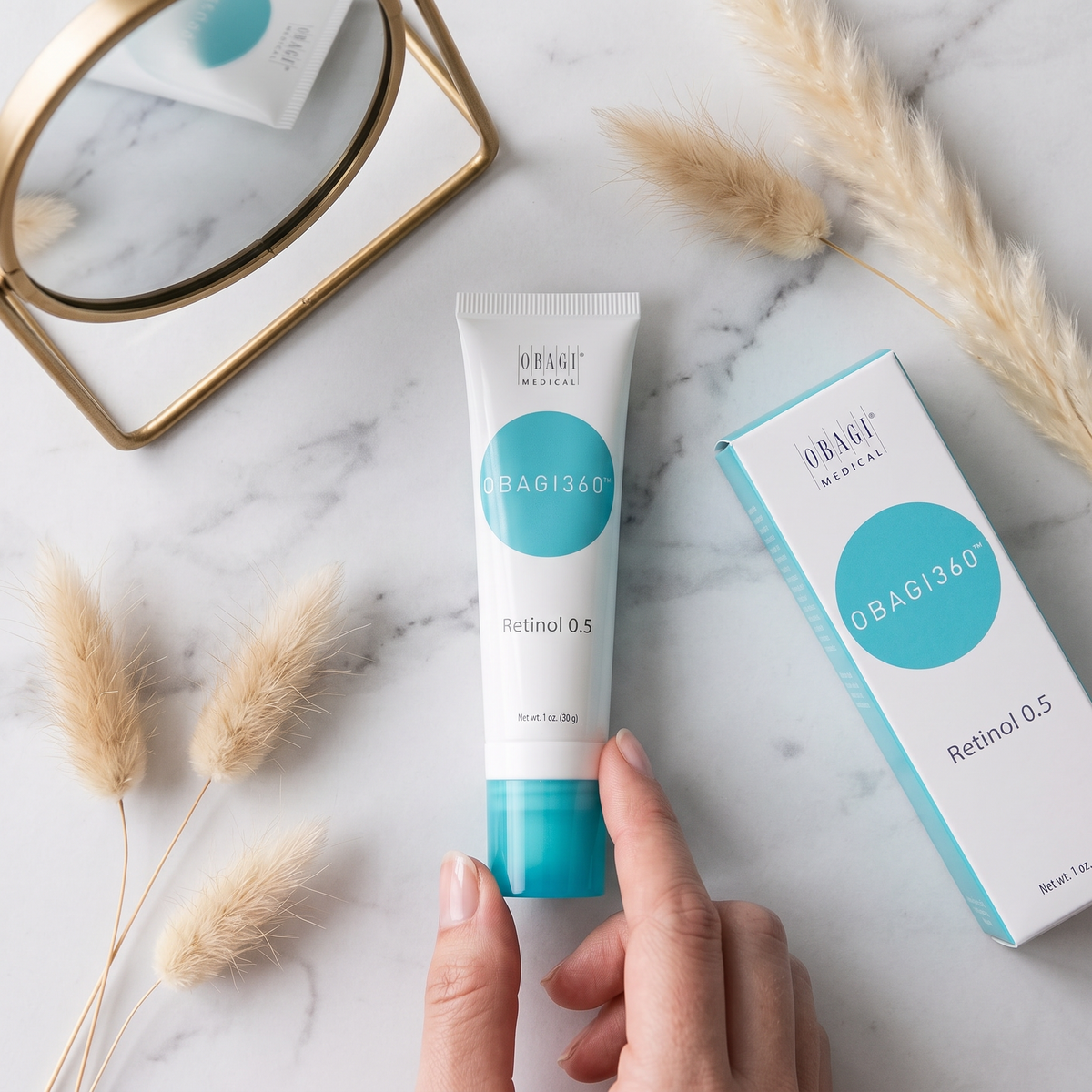 Elume Medspa Obagi360 Retinol 0.5 cream tube and box on a marble surface with dried grass and a mirror reflection.