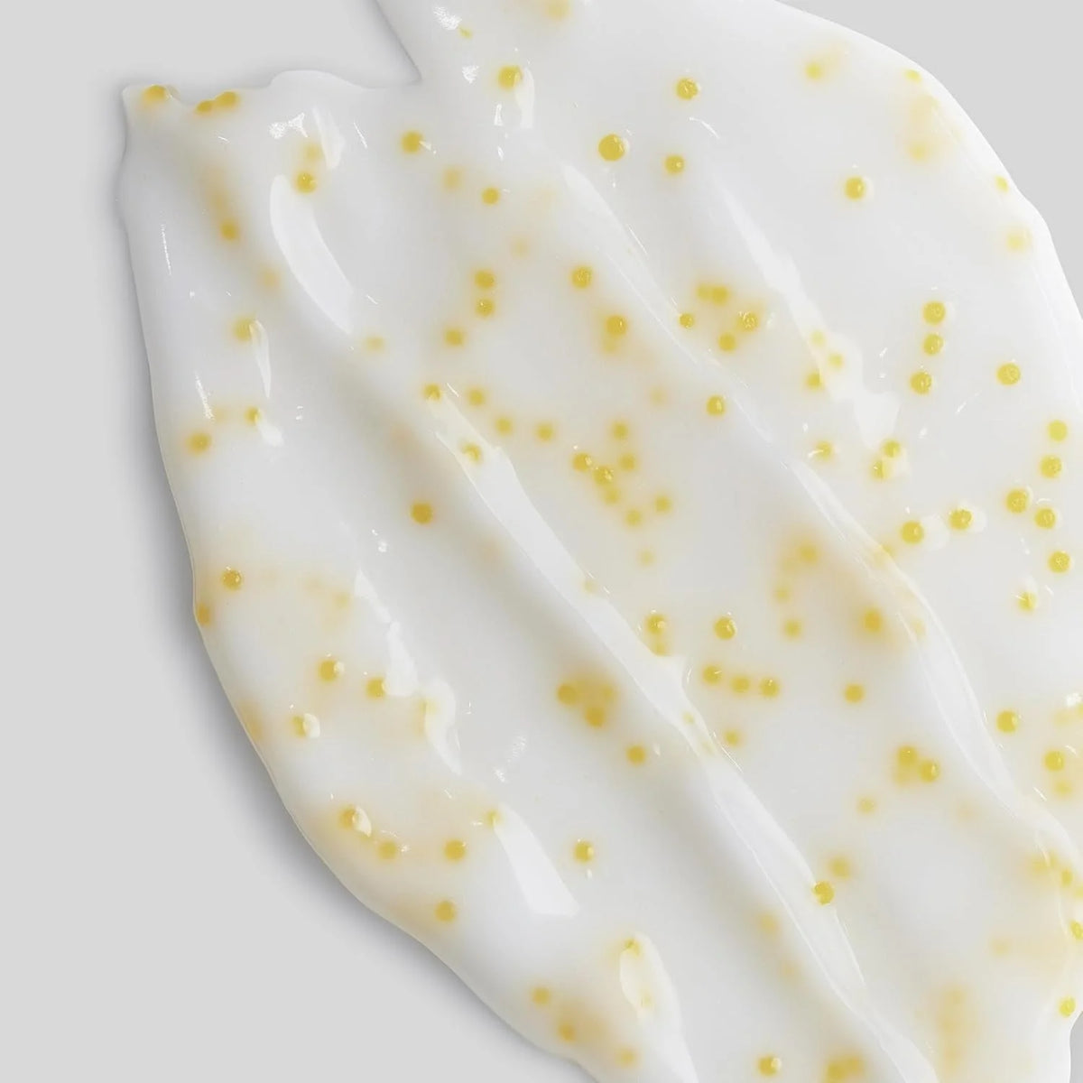 A close-up of a glossy white cream or gel swatch with scattered small yellow dots, photographed on a light gray background.